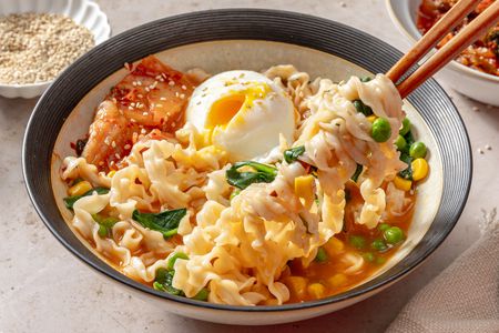 A bowl of noodle soup with an egg, kimchi, vegetables, and chopsticks lifting noodles, garnished with sesame seeds