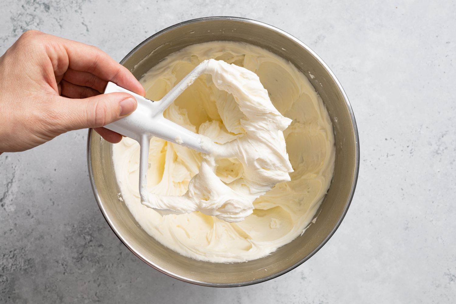 Hand Holding a Paddle Attachment with Icing for Lemon Lavender Cake. More Icing in Mixing Bowl 