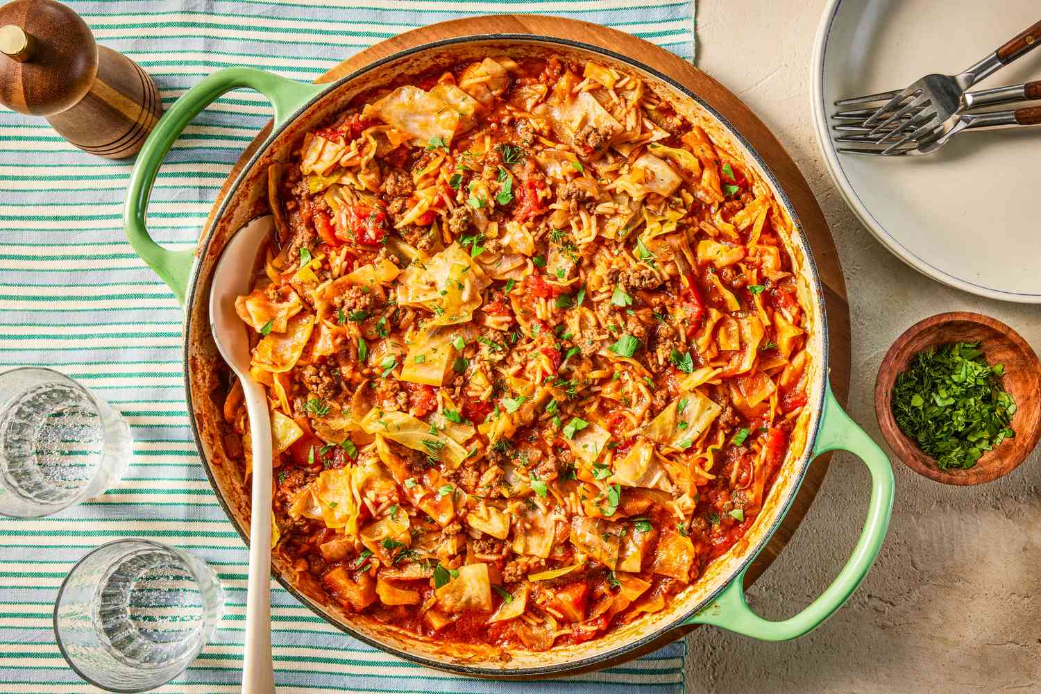 unstuffed cabbage rolls in a large pan at a table setting with a stack of plates, a small bowl of minced herbs, glasses of water, and a pepper cracker