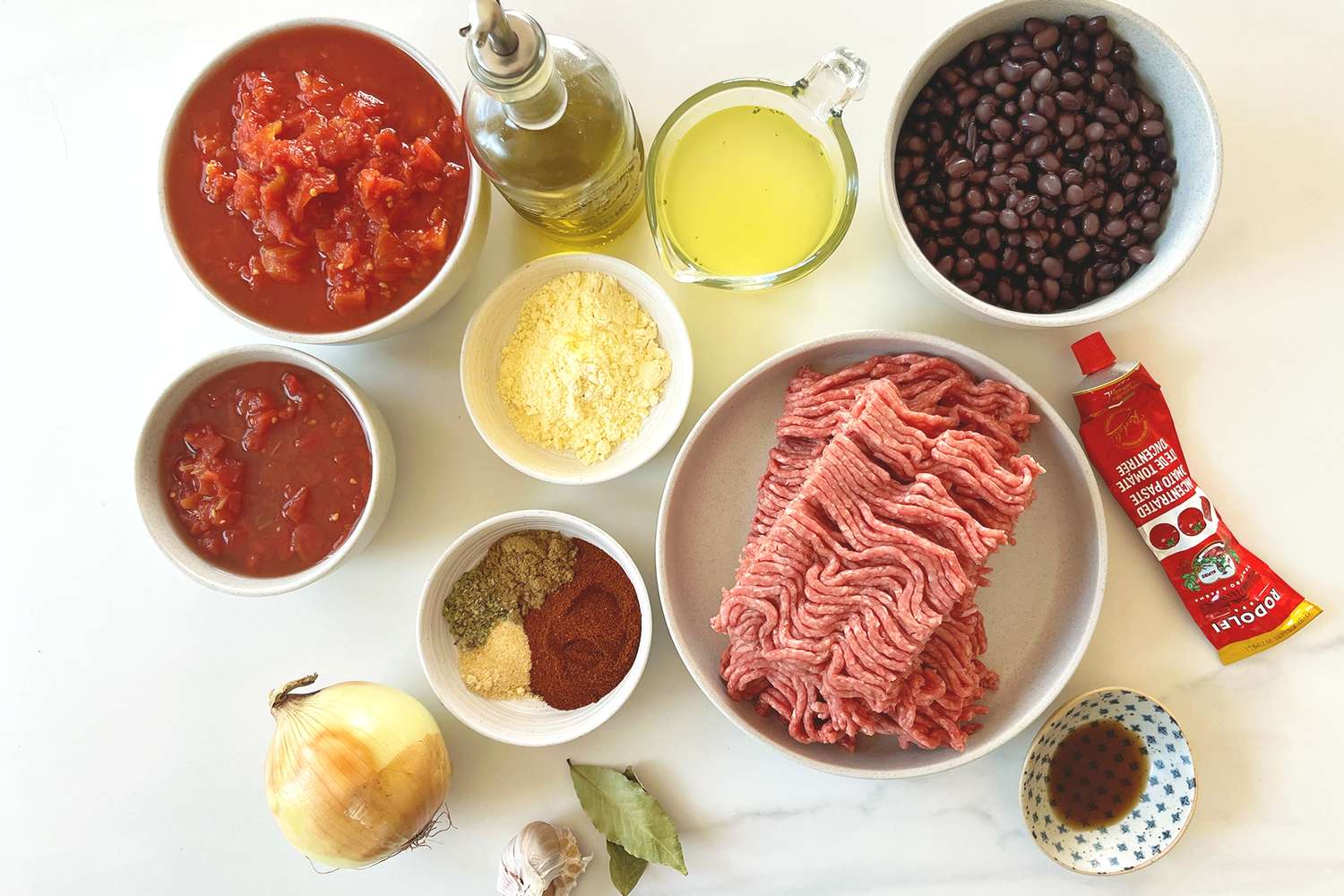 Ingredients for making chili displayed on a surface including ground meat black beans tomatoes spices and more