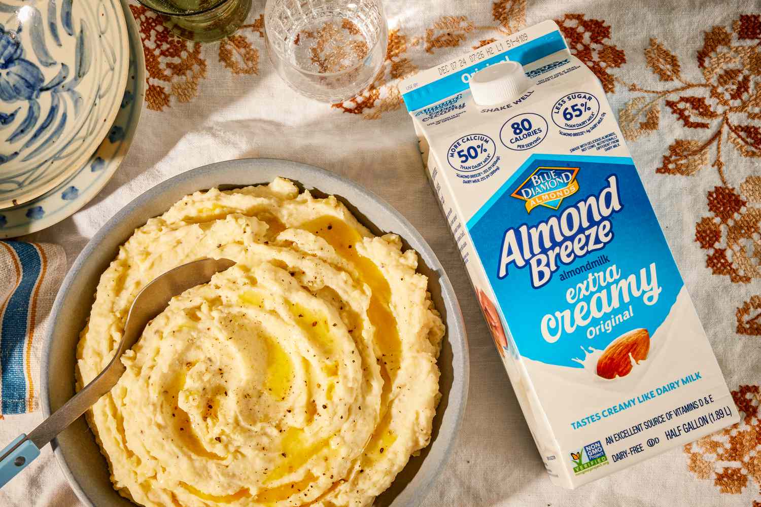 Overhead view of a gray bowl of almond milk mashed potatoes with a spoon and topped with butter next to a carton of extra creamy almond milk all on top of a fall themed embroidered cloth