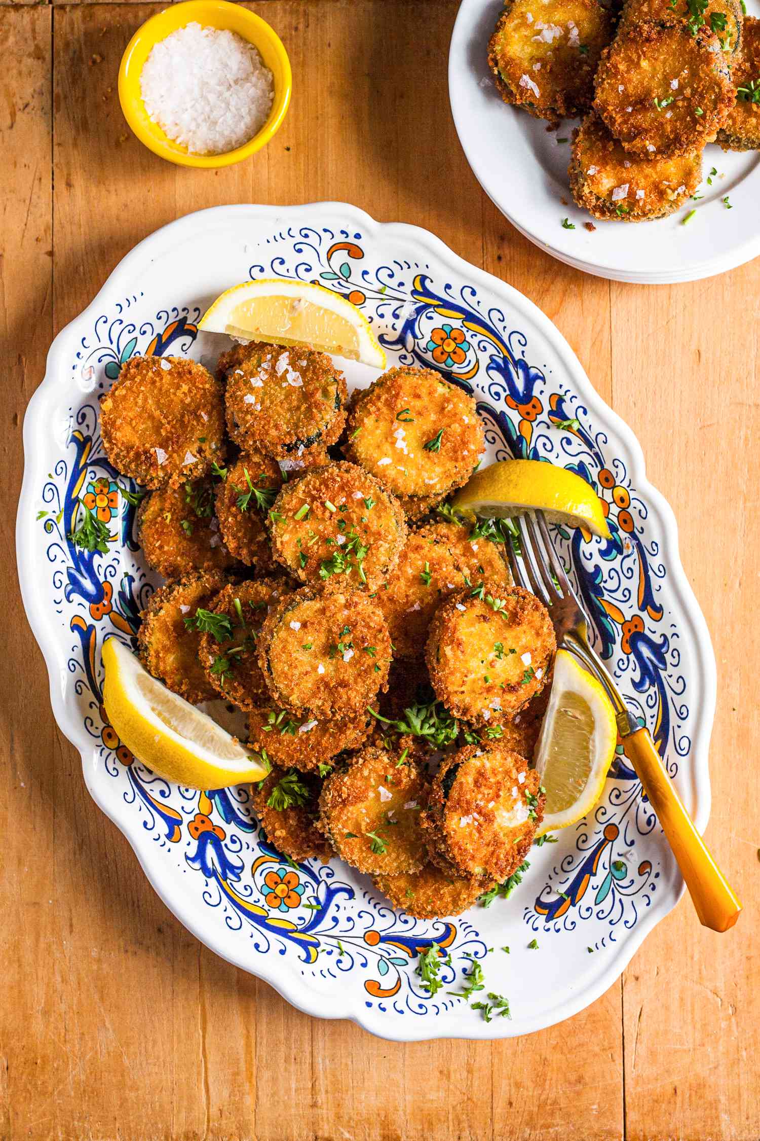 Fried Zucchini on a Platter with Lemon Wedges and a Fork, Next to a Small Bowl of Salt and Plate of Fried Zucchini