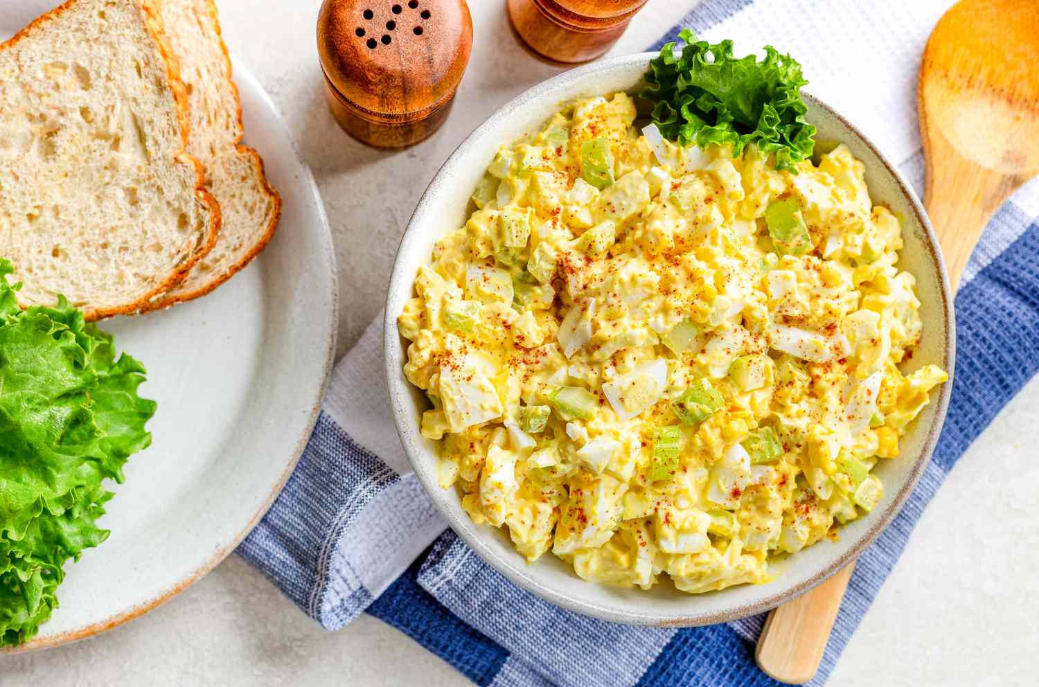 bowl of egg salad with a piece of lettuce on the counter next to a plate with bread and lettuce, salt and pepper shakers, and large spoon on a table napkin