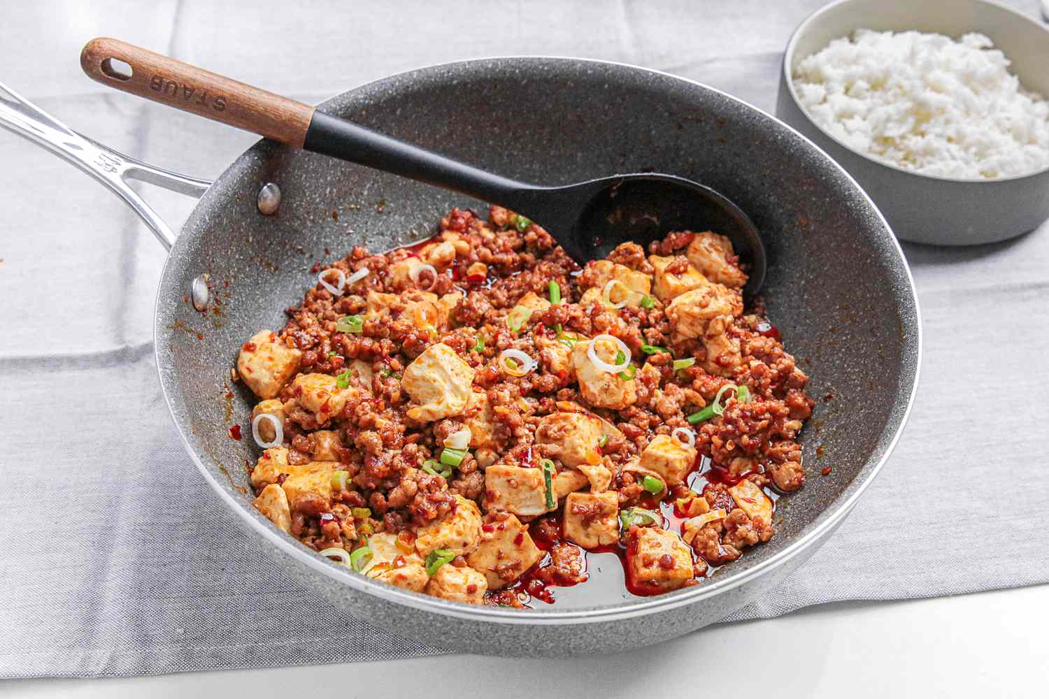 Mapo Tofu in a Pan With a Serving Spoon, and in the Background, a Bowl of Rice, All on a Grey Kitchen Towel