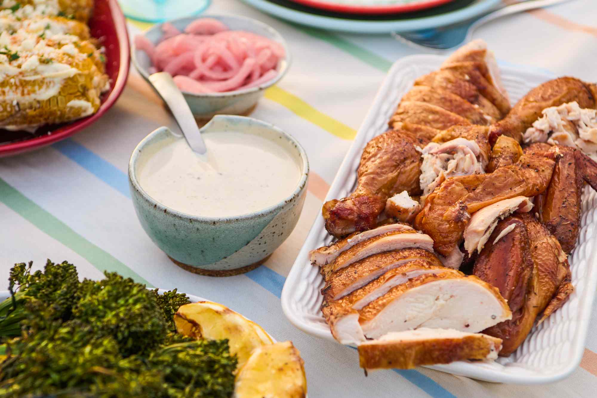 A platter of sliced grilled chicken with a bowl of white sauce and side dishes on a table