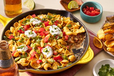 Frito pie dip in a cast iron skillet at a table setting with a bag of fritos lays, a single serving of dip on a plate, a small bowl of diced tomatoes, and a beer