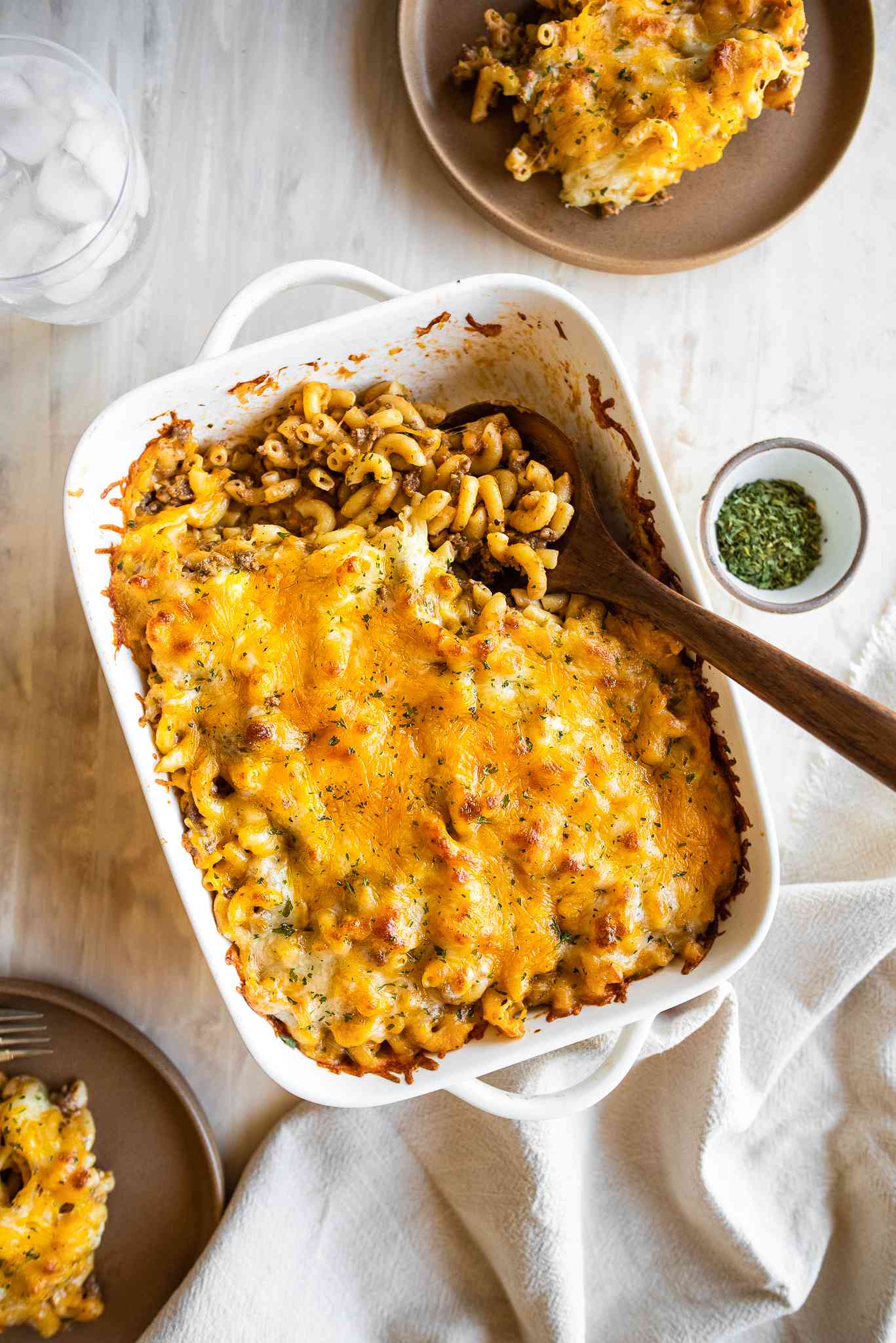Casserole Dish of Cheeseburger Casserole with a Serving Already Served Using a Spoon at a Table Setting (a Plate with a Serving of Casserole, Bowl of Dry Parsley, a Table Napkin, and a Glass of Ice Water)