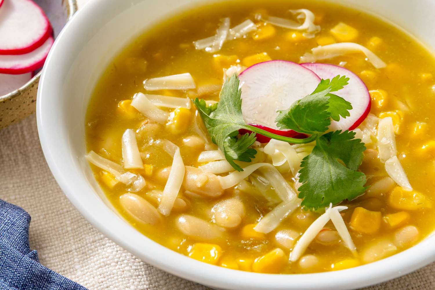 A bowl of chili verde topped with radish slices and cilantro garnished with shredded cheese and corn