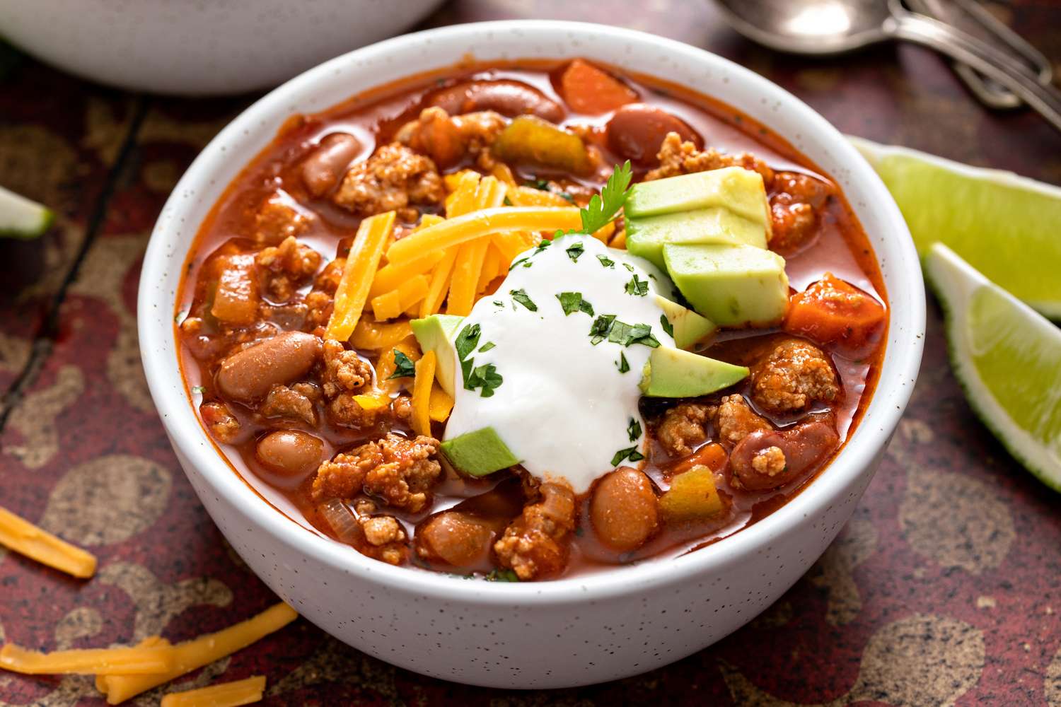 A bowl of chili topped with sour cream shredded cheese and avocado slices accompanied by lime wedges on the side