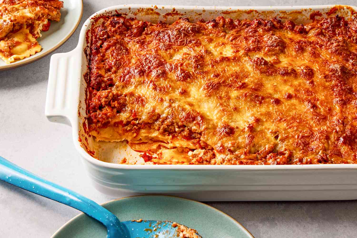 Side view of a white baking dish of Million Dollar Ravioli Casserole with a serving removed and on a small plate next to a serving spoon on a small plate