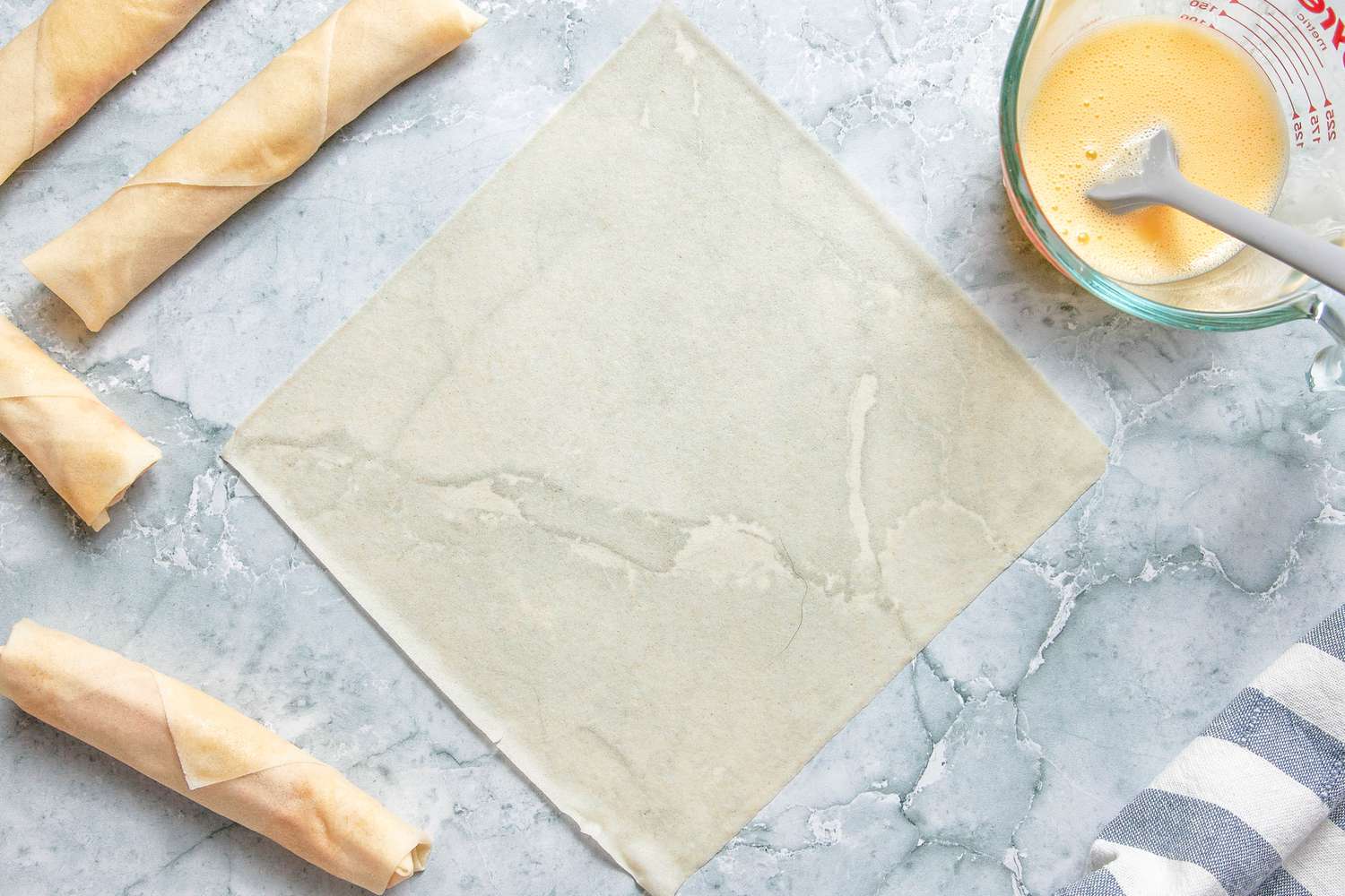 Egg Roll on the Counter (Positioned Like a Diamond), and in the Surroundings, Rolled Lumpia on the Counter, a Pyrex Measuring Cup With the Egg Wash, and a Kitchen Towel Also on the Counter