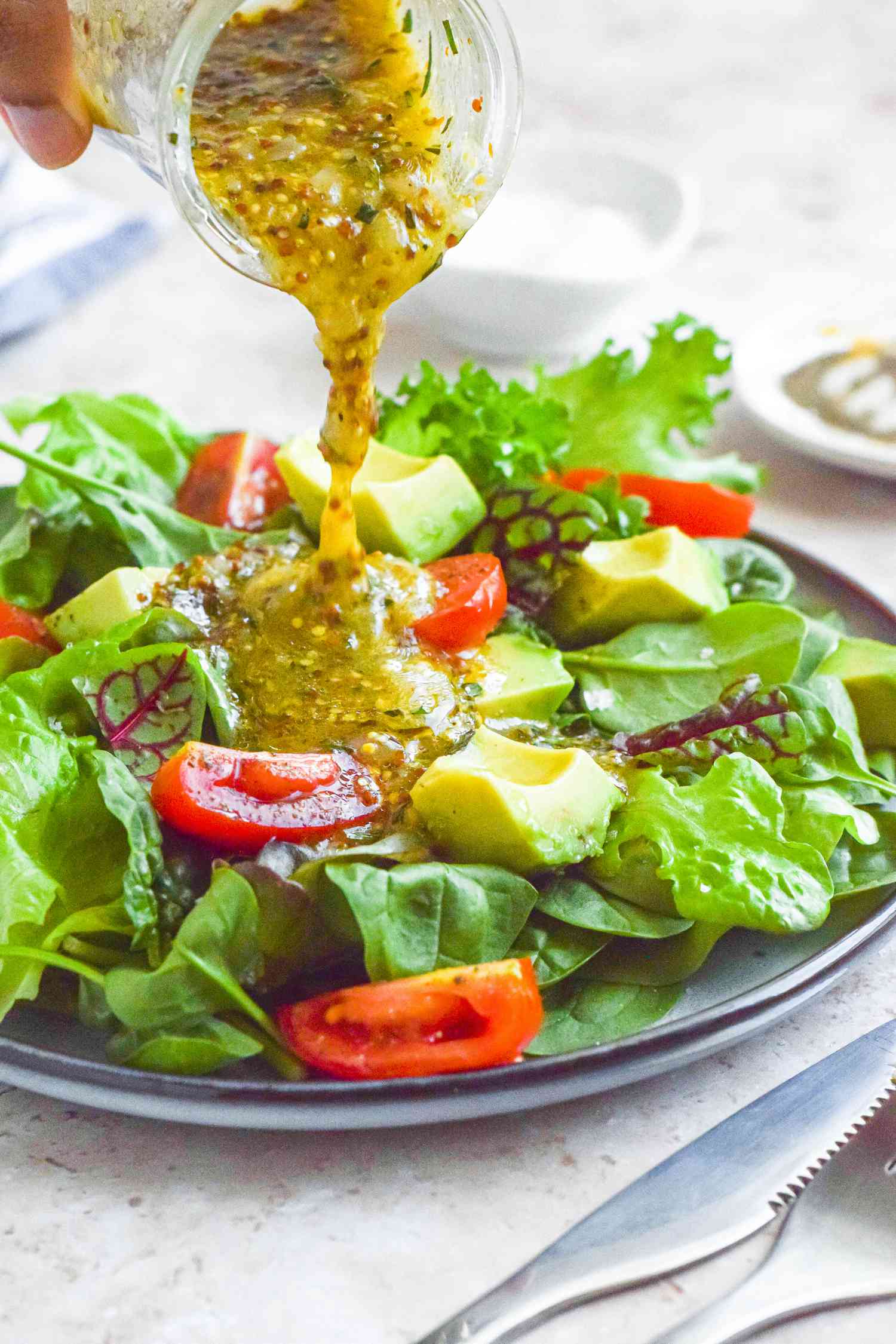 Jar of Tarragon-Mustard Vinaigrette Poured onto a Salad (Various Lettuce Types, Sliced Tomatoes, and Sliced Avocado)