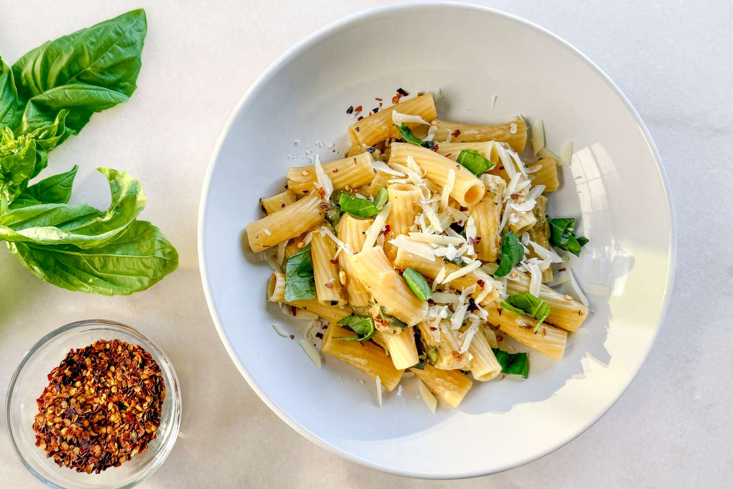 Overhead shot of a white shallow bowl withe rigatoni pasta, topped with shredded cheese and basil