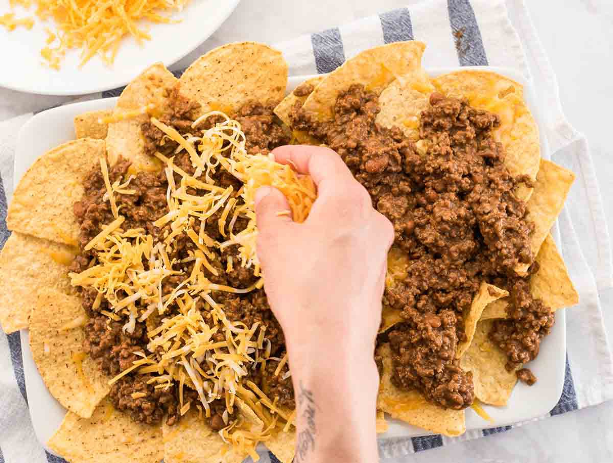 Shredded cheese being sprinkled on to ground beef covered tortilla chips on a white platter