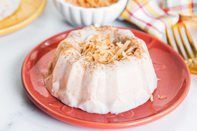 Homemade bundt shaped templeque is on a coral plate with a gold spoon set to the left. The tembleque has toasted, shredded coconut mounded in the center. Behind the plate is a striped dish towel, small white container and yellowl plate all in partial view.