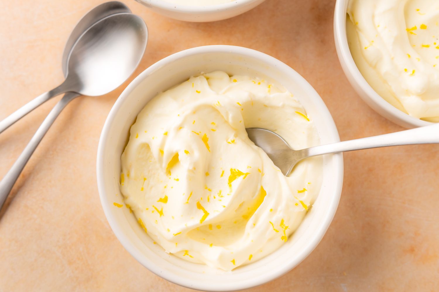 Overhead view of small white bowls of lemon mousse next to a spoon and plate of lemon slices all on an orange surface