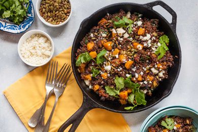 Horizontal view of a large cast iron skillet filled with a butternut squash skillet dinner. Cilantro, quinoa, butternut squash and cotija cheese are visible in the pan. To the left is a yellow napkin with three forks on it. Above the forks are three small bowls. One small bowl of rice, a ramiken of roasted pumpkin seeds and a small platter of cilantro.