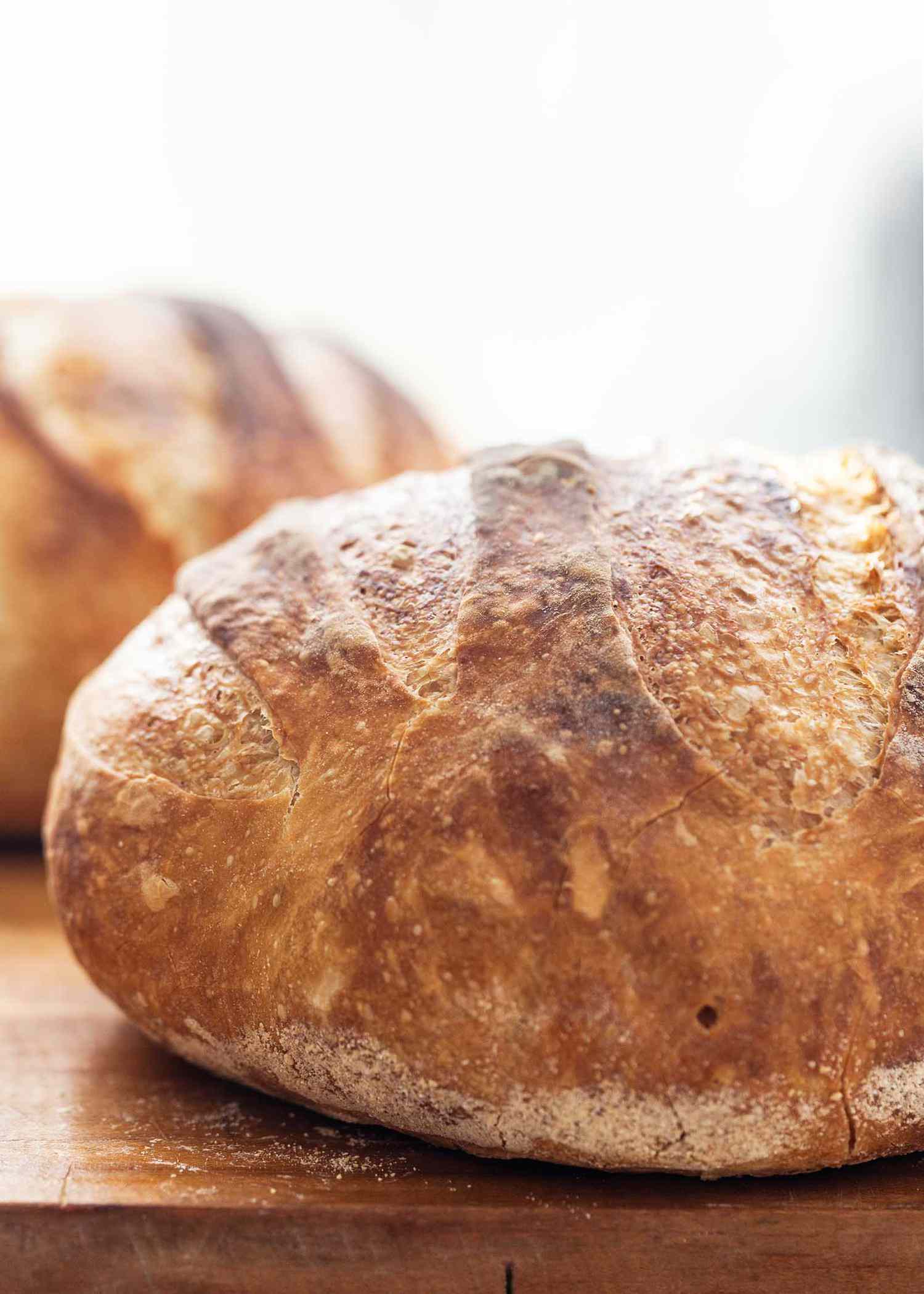 Side view of no-knead dutch oven bread on a cutting board. The crust is deep golden brown and a second loaf is behind the first.