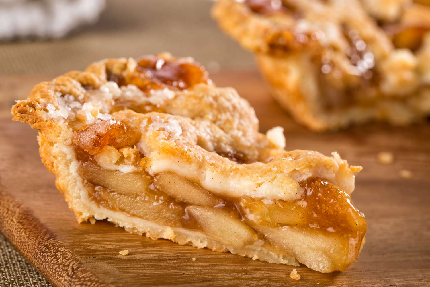 Close up side view of a slice of apple pie with the full pie blurred in the background all of a bamboo cutting board