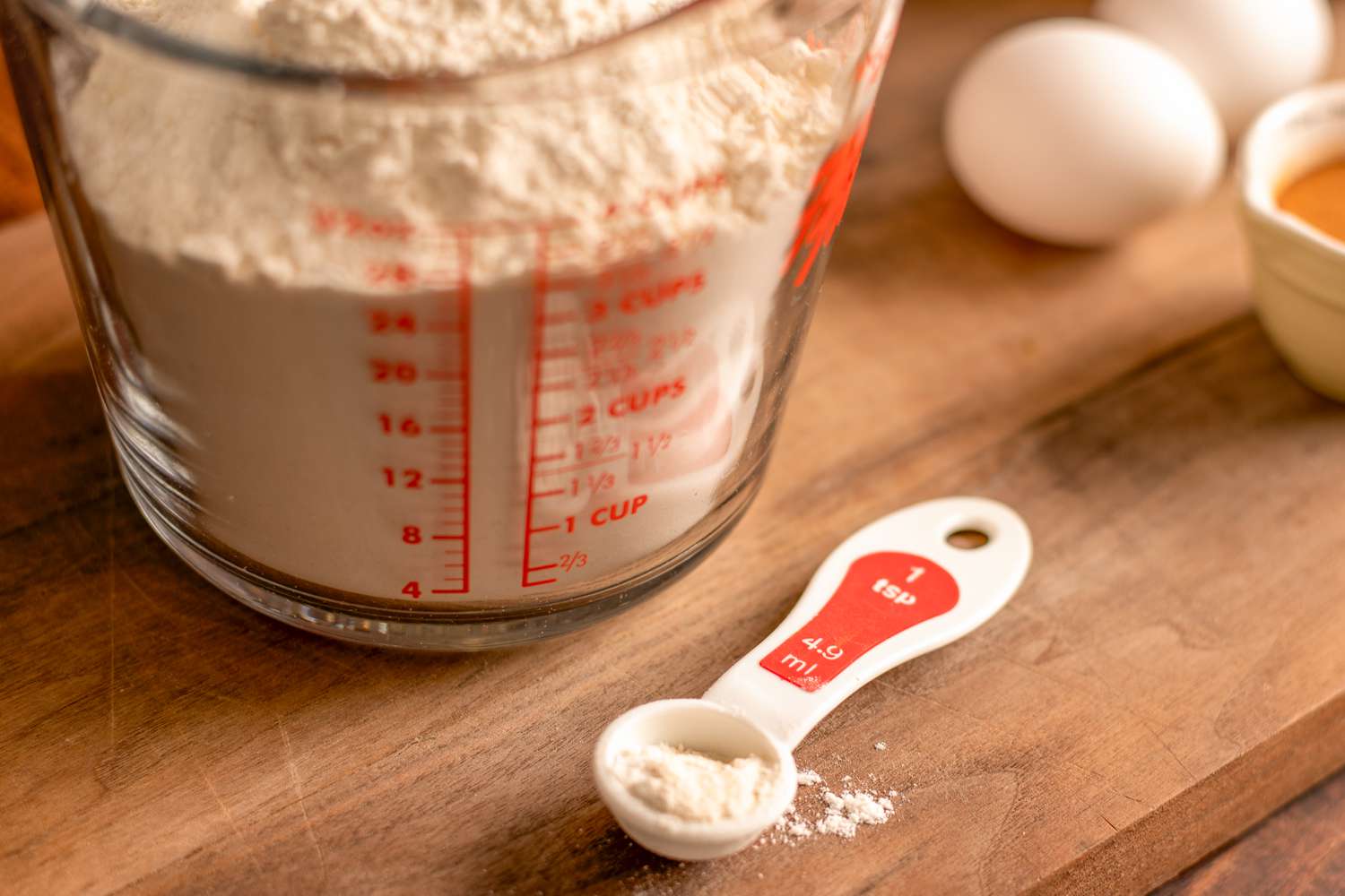 A glass measuring cup filled with flour next to a teaspoon with white powder in it; two eggs are in the background