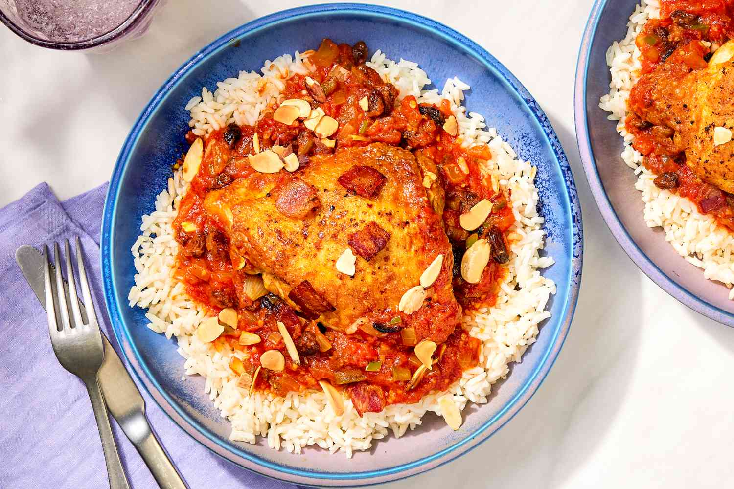 Overhead view of two blue bowls of Country Captain Chicken served over a bed of rice next to a fork, knife and cloth napkin