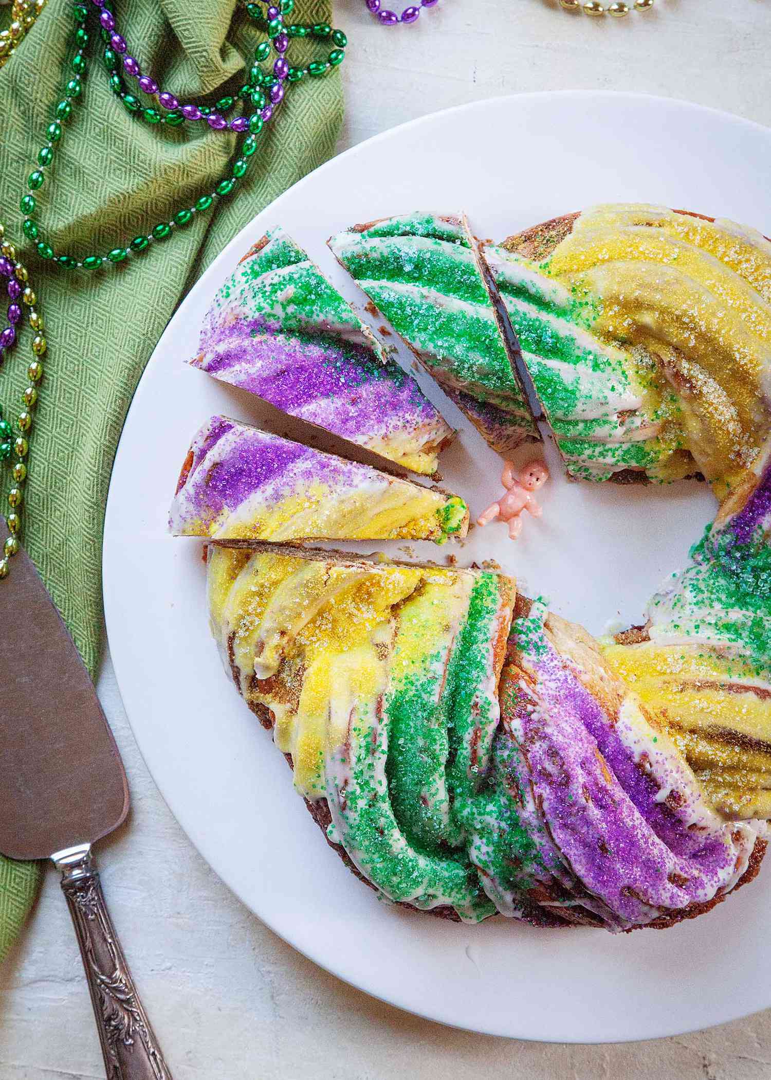Top view of the best king cake cut into slices and resting on a white platter. A metal serving utensil, Mardi Gras beads and a green linen are to the left.