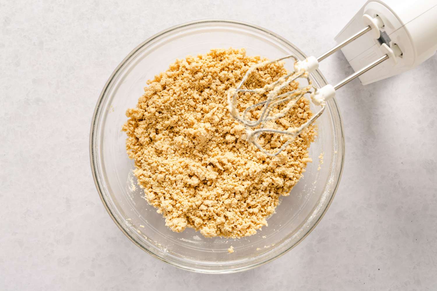 A glass bowl containing crumbly ingredients for caramel apple bars recipe with a handheld electric mixer nearby