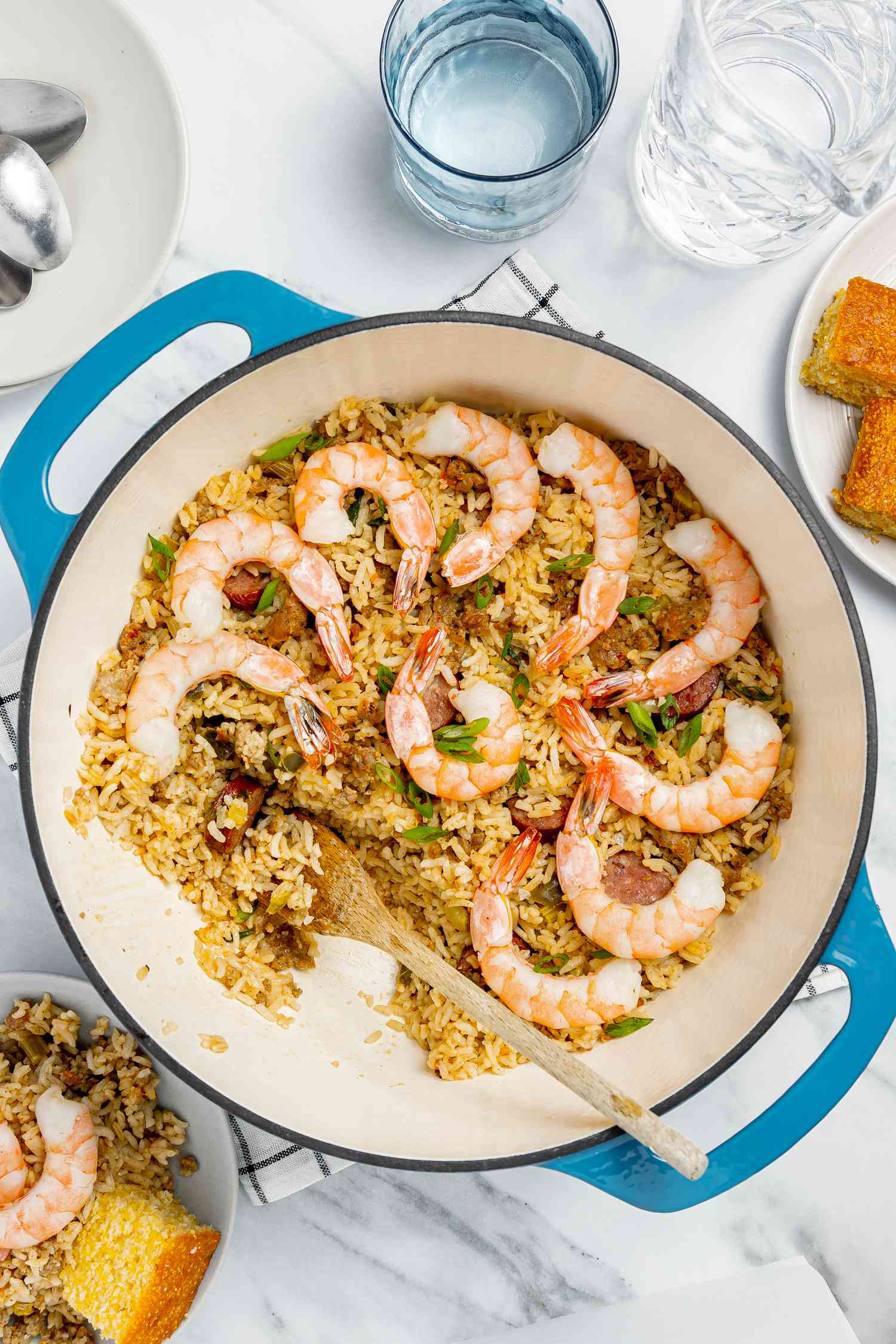 Dirty Rice With Shrimp in a Dutch Oven, and Next to It a Bowl With a Serving of Rice and a Piece of Cornbread, Glasses of Water, a Plate With Utensils, and a Plate With Cornbread 