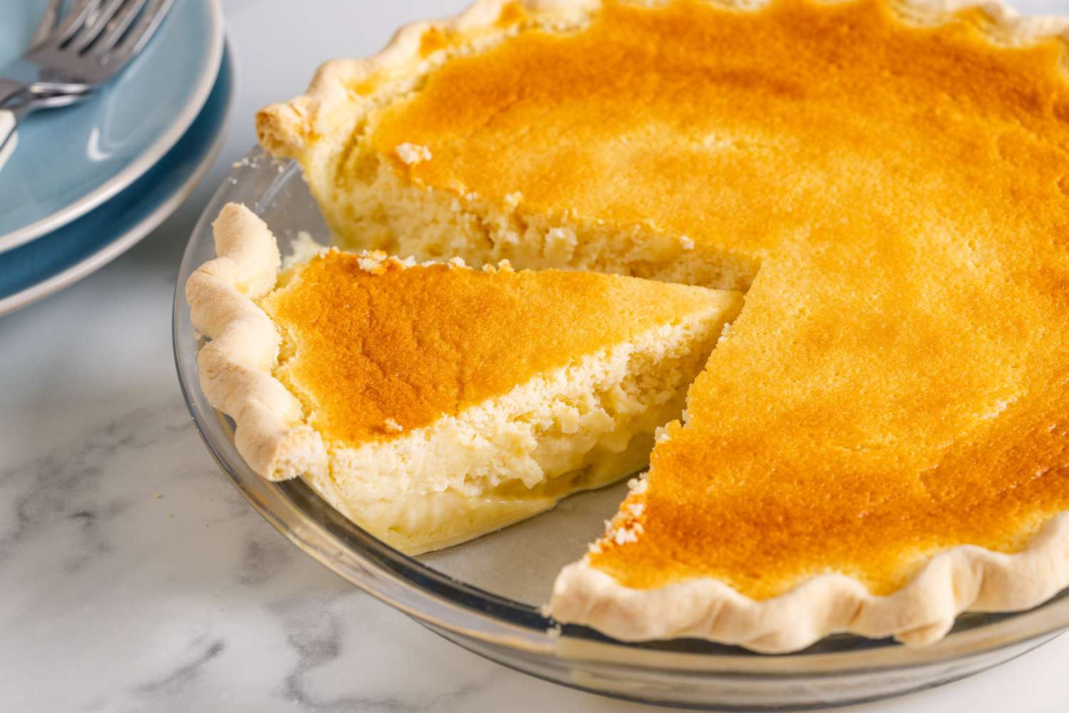 A lemon pie with a slice cut out displayed in a glass pie dish on a marble surface
