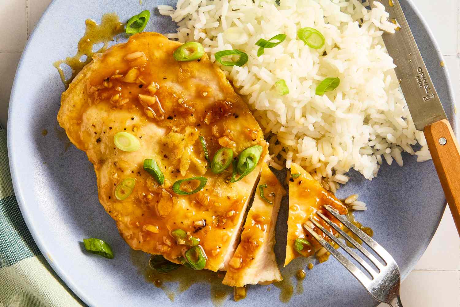 Overhead view of a blue plate with a serving of partially sliced honey soy chicken and white rice topped with green onions along with a fork and knife