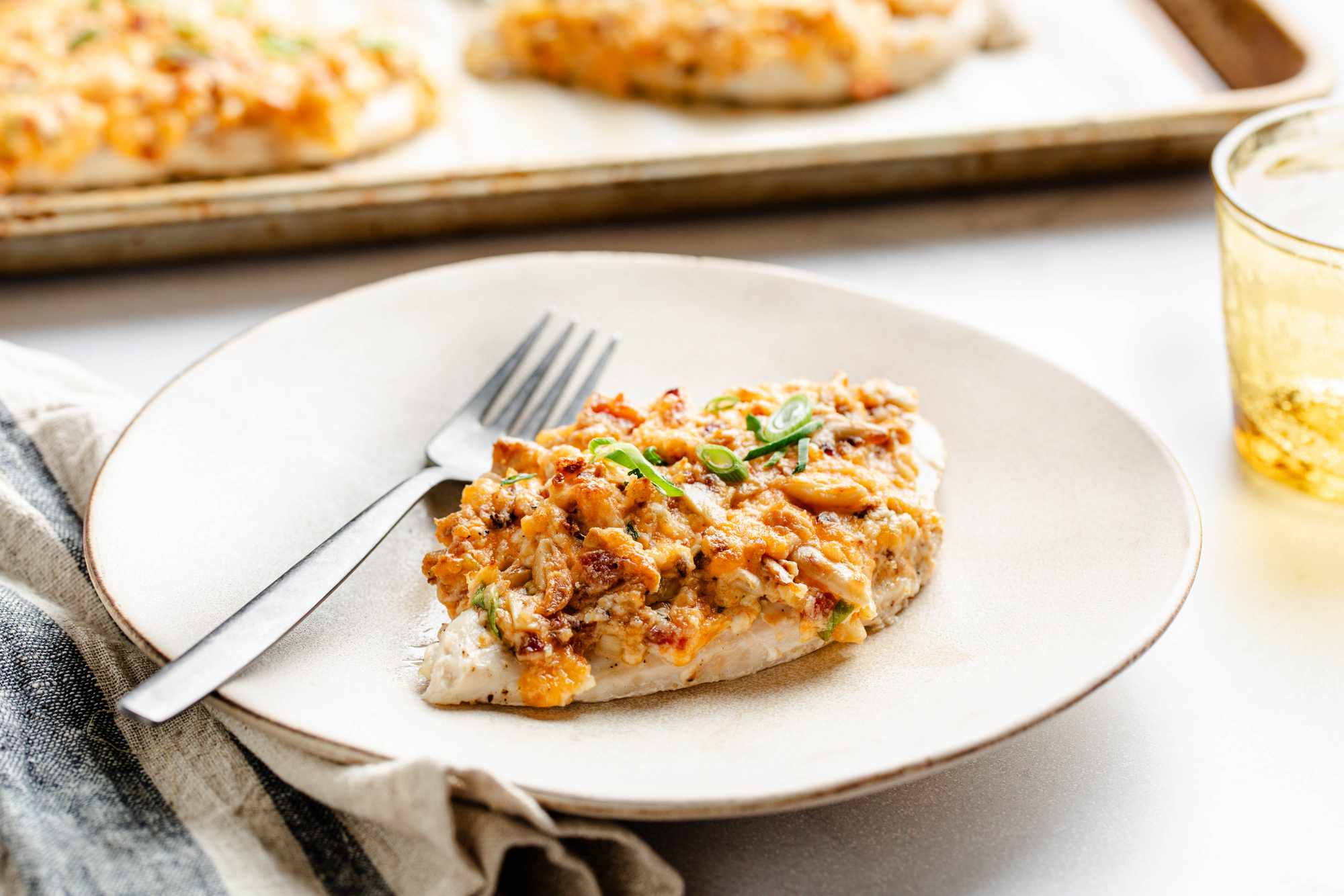 A million dollar chicken breast on a plate with a fork, and in the surroundings, a parchment paper lined baking tray with more, a glass of water, and a table napkin