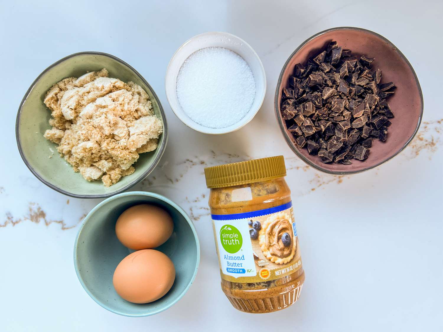 Four bowls filled with cookie ingredient next to one jar of almond butter.