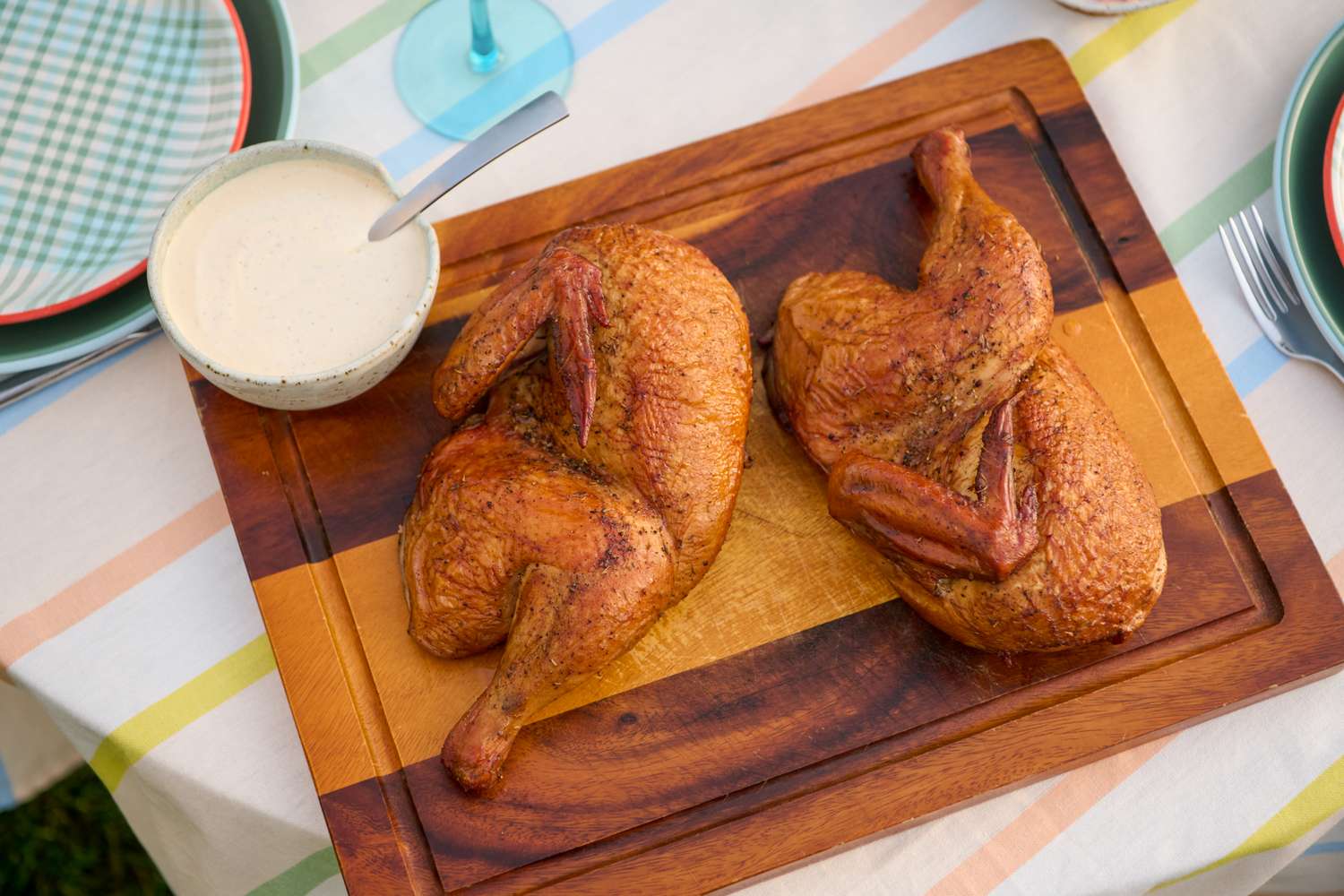 Two grilled half chickens on a wooden serving board, accompanied by a bowl of sauce with a spoon