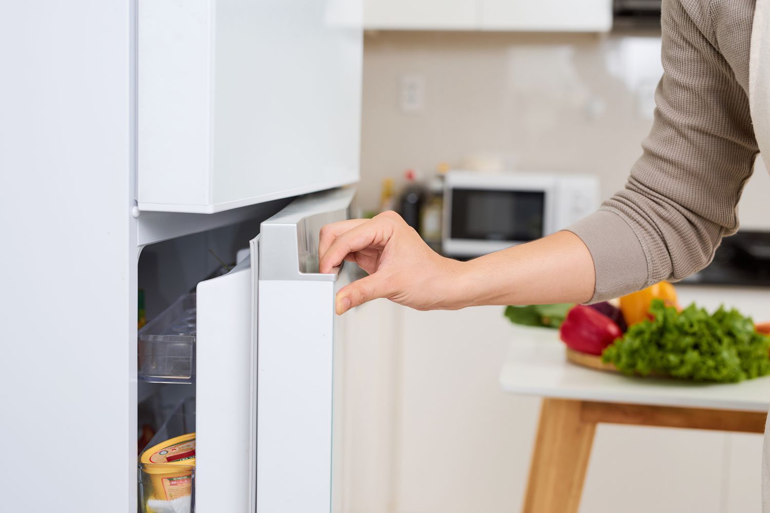 A young man opening a refrigerator door in a kitchen with a microwave visible in the background