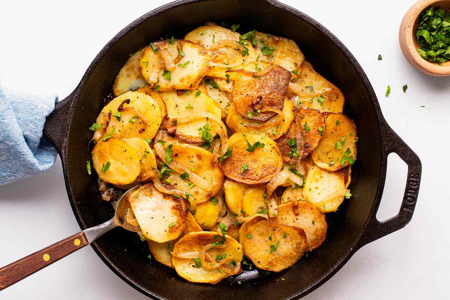 Lyonniase potatoes in a cast iron skillet with a spoon and next to a bowl of minced parsley