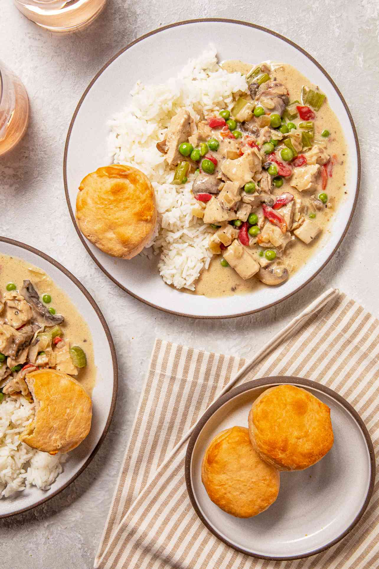 Chicken a la King on a plate with rice and a biscuit at a table setting with another plate of chicken a la king, a small bowl of biscuits, glasses of wine, and a table napkin