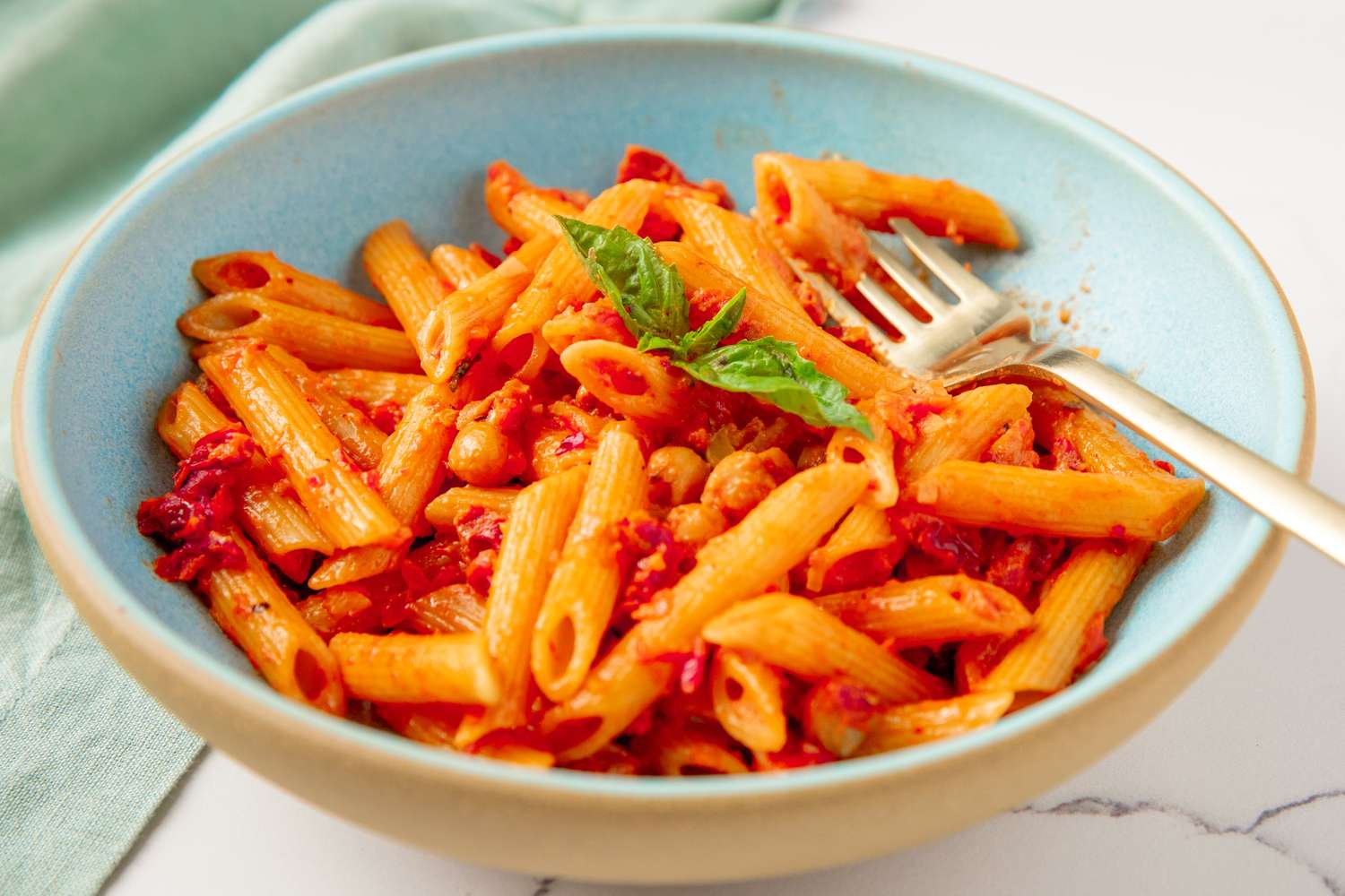 A bowl of penne pasta with chickpeas and marinara sauce, garnished with a basil leaf and a fork rested on the side