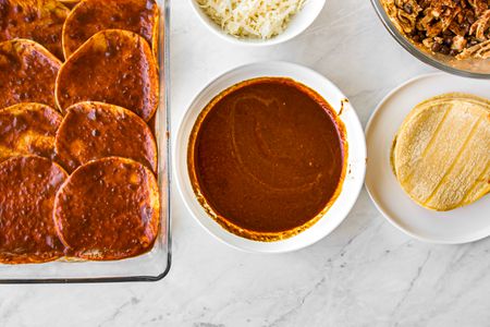 Bowl of Red Enchilada Sauce Surrounded by Enchilada Ingredients (a Plate of Tortillas, Bowl of Shredded Chicken, Bowl of Shredded Cheese, and Glass Platter with Sauce Soaked Tortillas)