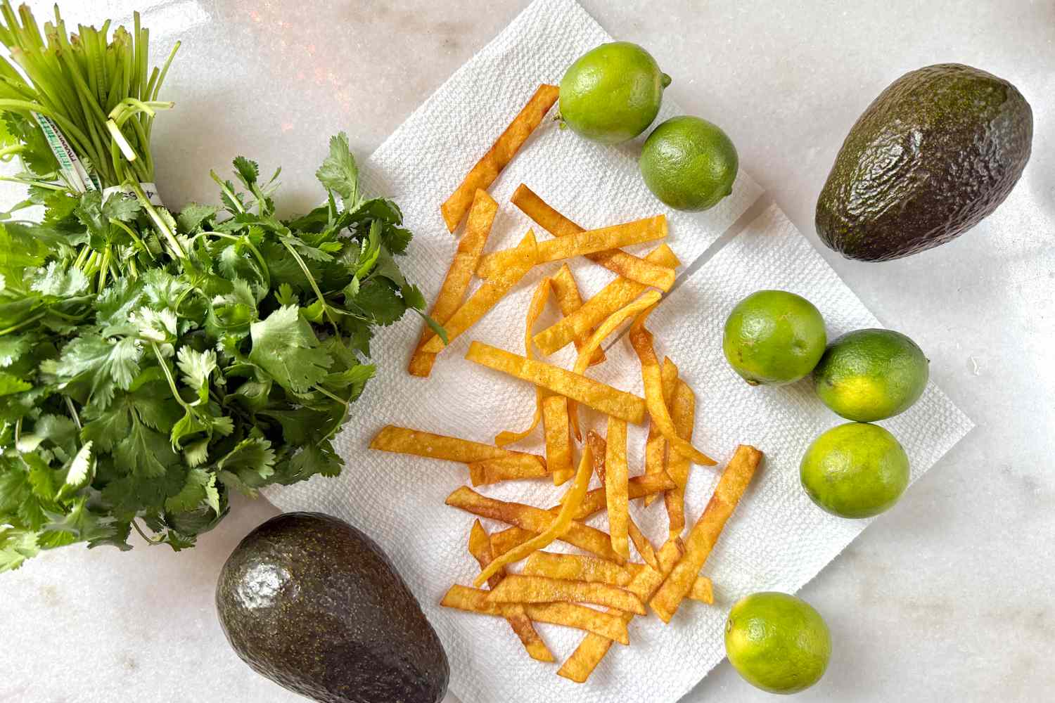 Overhead view of tortilla strips, limes, avocados and fresh cilantro on a white countertop