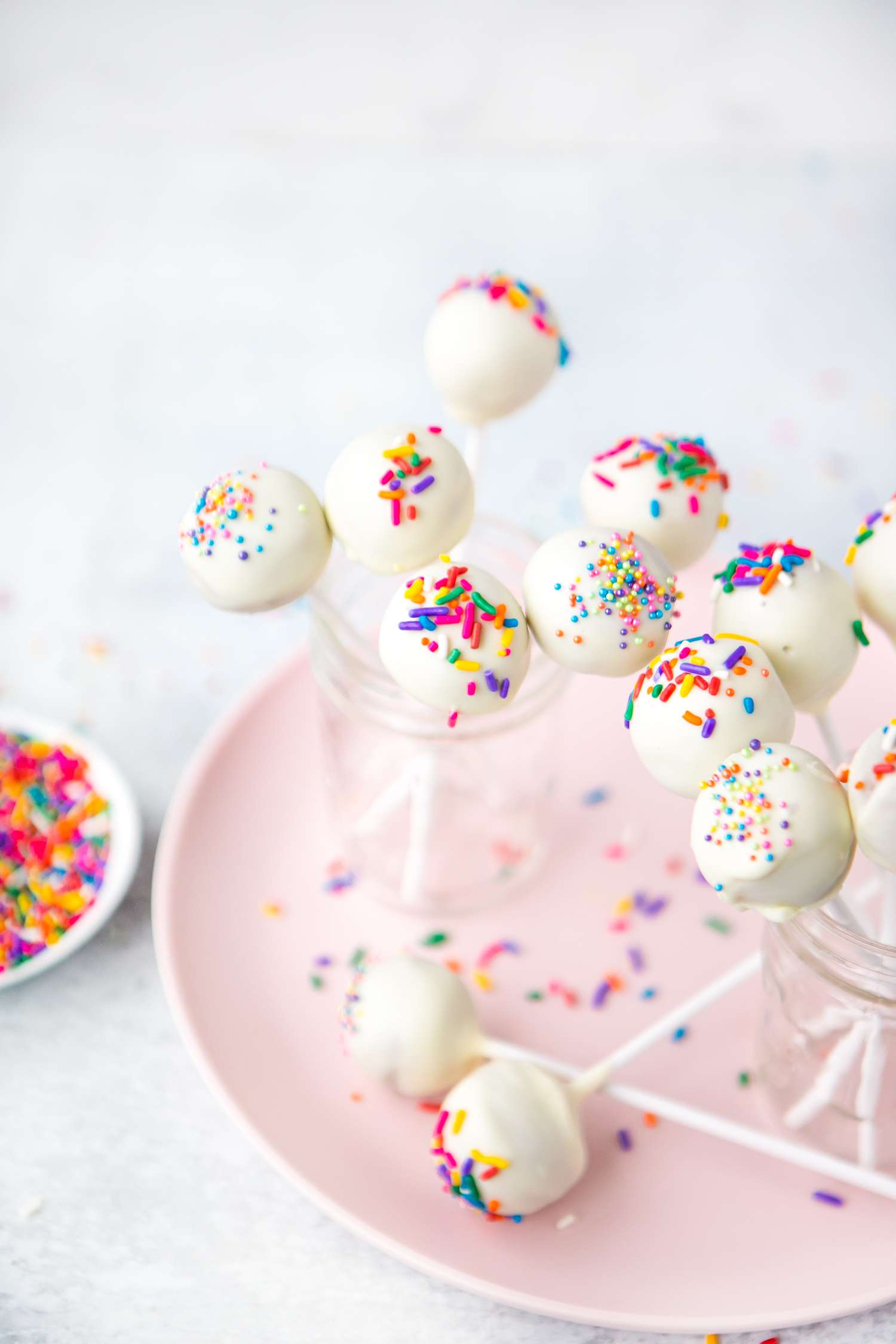 Sprinkled Cake Pops in Two Jars on a Plate Next to Small Plate of Sprinkles