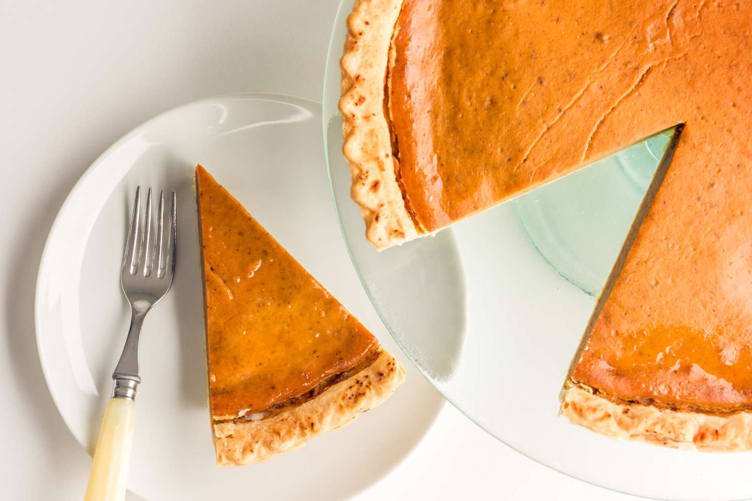 Pumpkin pie on a glass serving platter, and a slice of pie on the plate next to it