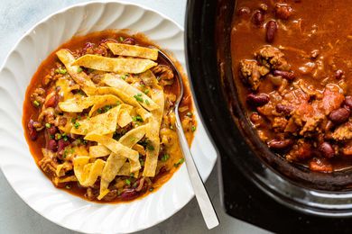 A bowl of beef and bean chili that's garnished with fried tortilla strips and sitting next to a slow cooker