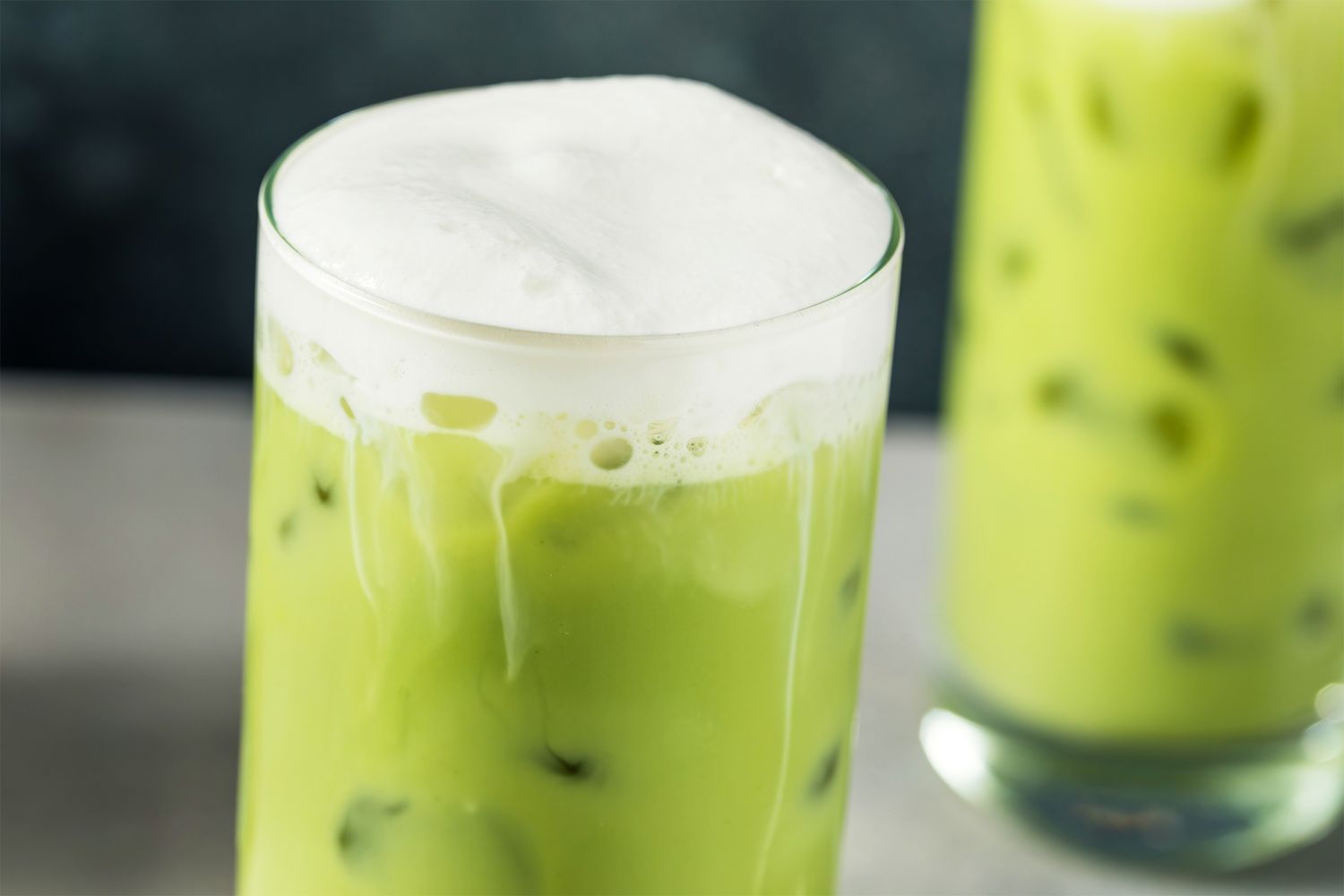 A glass of iced matcha latte topped with foam with another glass in the background