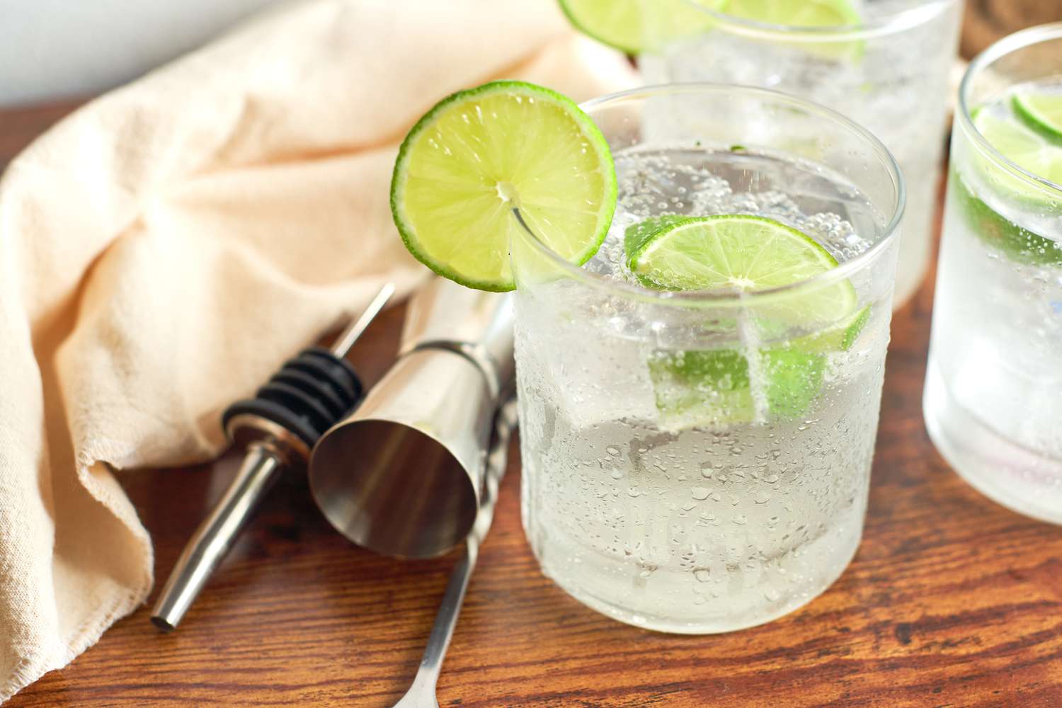 Side view of three glasses of tonic water with lime pieces on a wooden tabletop next to a napkin and bar equipment