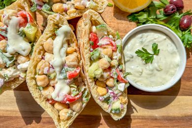 Greek salad pockets next to a some olive, fresh herbs, a halved lemon, and small bowl of tzatziki, all on a cutting board 