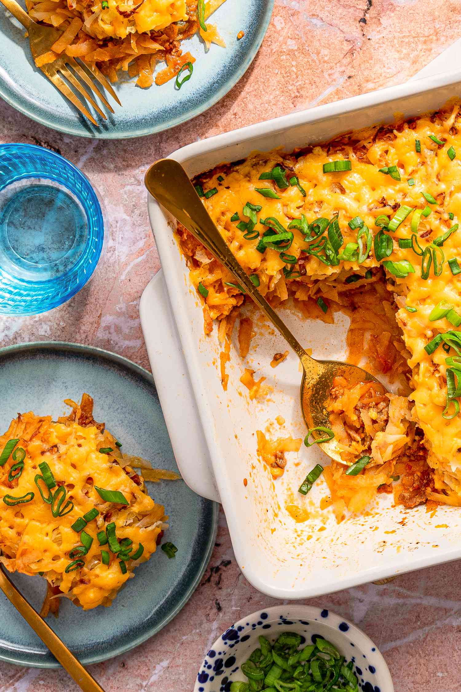 shipwreck casserole in a casserole dish and a few servings on a couple of plates at a table setting with a glass of water and a bowl of sliced scallions
