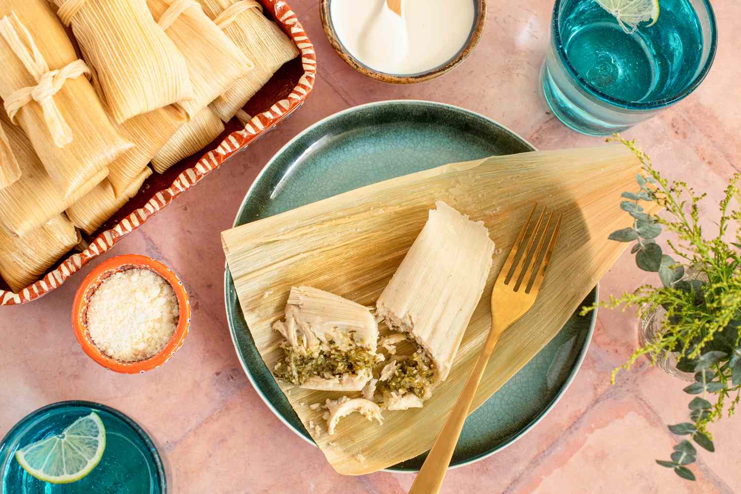 A Plate of Chicken Tamales with Chile Verde Broken in Half to Show the Filling at a Table Setting with a Platter of More Tamales, Utensils, a Small Bowl of Cotija Cheese, a Small Bowl of Crema, a Glass of Water, and a Small Vase