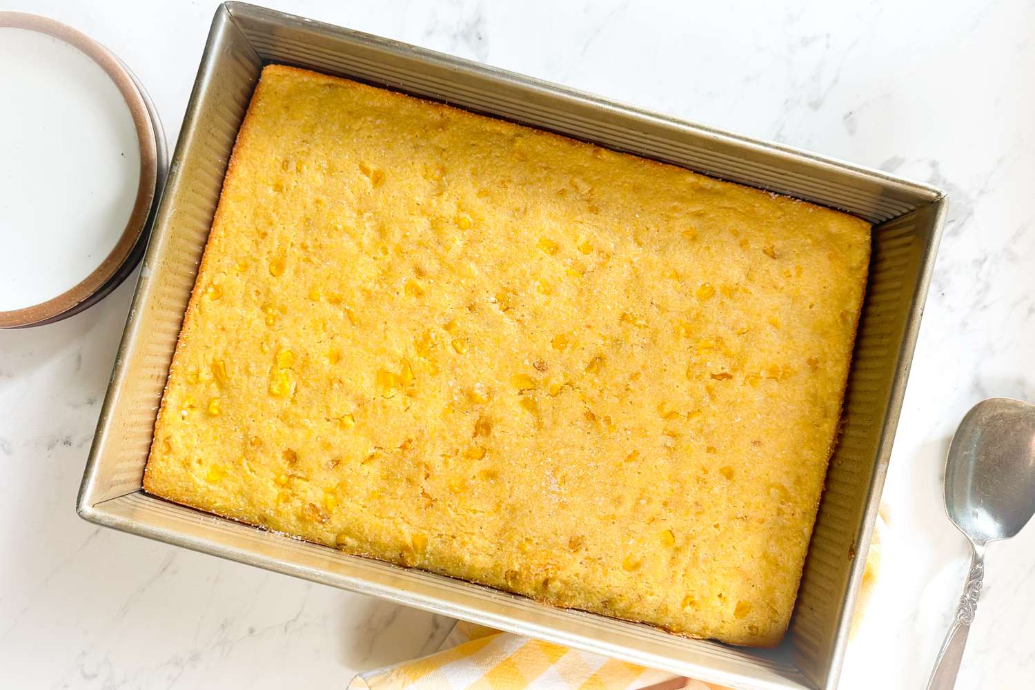 Baked cornbread in a metal pan on a surface with a spoon nearby