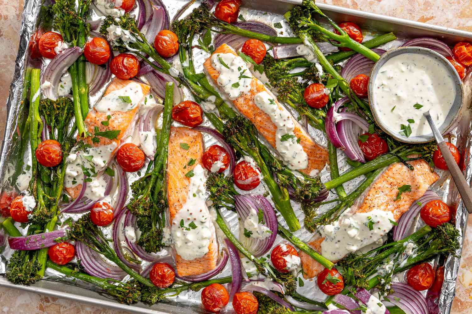 Overhead view of a sheet pan of salmon, vegetables and feta sauce with a bowl of sauce with a spoon