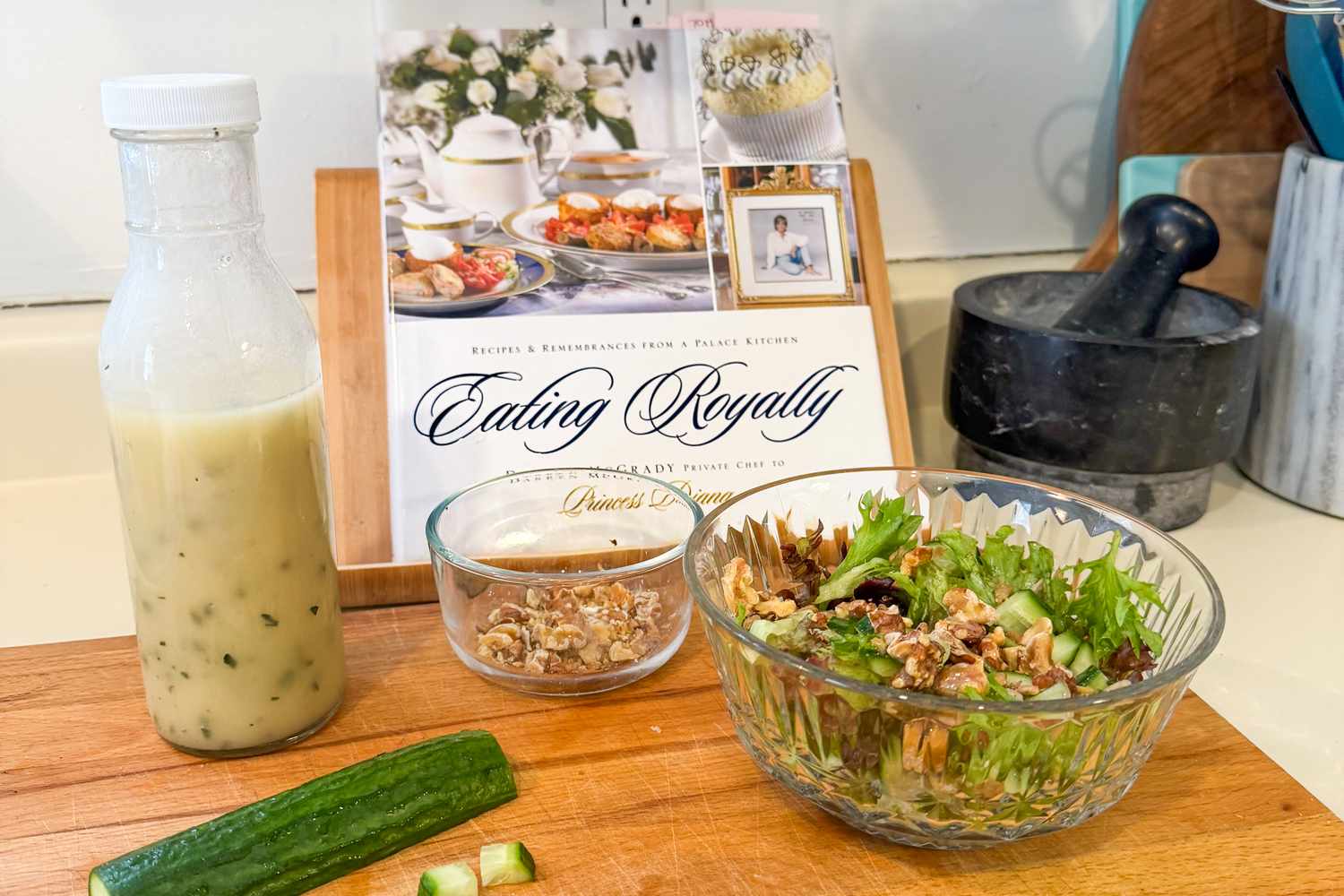 A salad with walnuts and a glass bottle of dressing on a wooden surface with an Eating Royally cookbook in the background